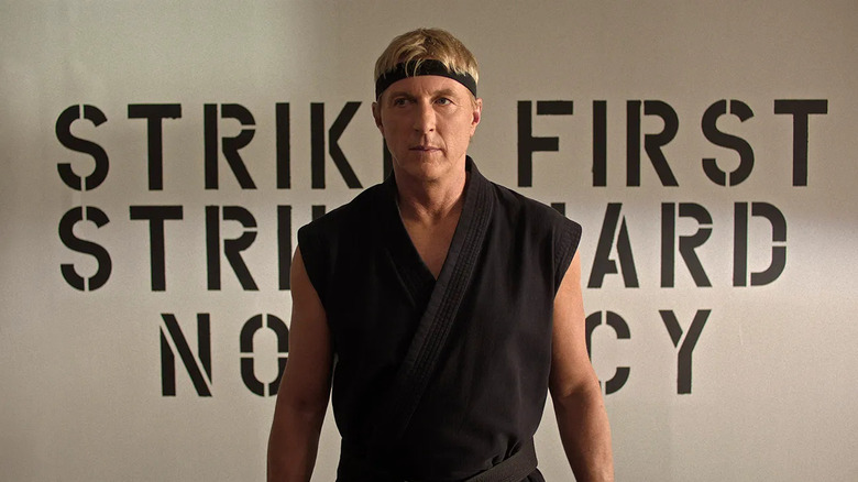 William Zabka as Johnny Lawrence, standing in front of the Cobra Kai mantra on Cobra Kai